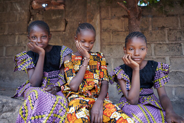Three beautiful young welldressed black African girls sitting side by side in front of a wall with...
