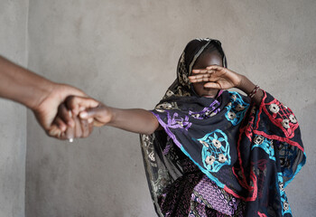 Veiled black African girl with her hand covering her eyes, pulled by the hand of an adult towards...