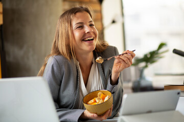 Businesswoman in office having healthy snack. Young woman eating fruit while having a video call..