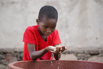 Little black African boy fetching water with his cupped hands from a tub to wash his face or to...