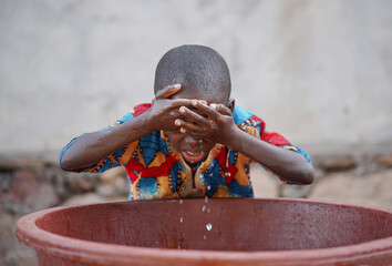 Little black African toddler rubbing his forehead with fresh and clean water from a plastic basin...