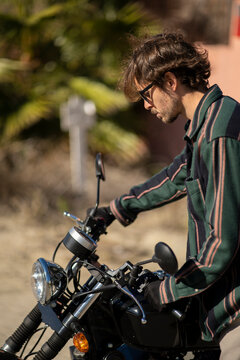 Adolescente hombre joven motero con camisa de cuadros elegante prepar&aacute;ndose para viaje. Fondo de palmeras veraniegas. Chico elegante con gafas de sol y moto. Chico poni&eacute;ndose casco de seguridad.