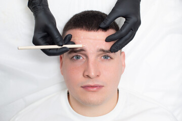 Portrait of a young handsome man with a wrinkled forehead, while the hands of a cosmetologist in black medical gloves hold a white cosmetic pencil and make marks on the guy face