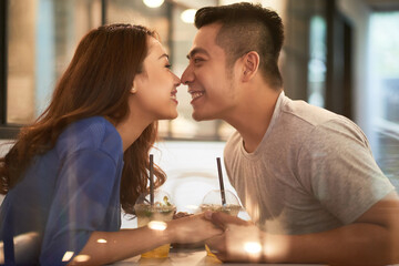 Happy affectionate young Asian couple sitting with summer cocktails and touching noses above table in cafe