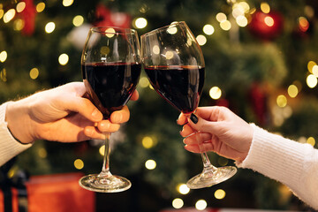 Close up Hands of unknown people who cheering with glasses of wine against Christmas tree decorated with glowing garland. Blurred background, bokeh. Happy New Year