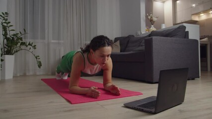 Adult woman in sportswear performing abs workout, doing yoga plank exercise, watching online video tutorial on laptop, getting exhausted, falling on mat floor indoors. Training for weight loss at home