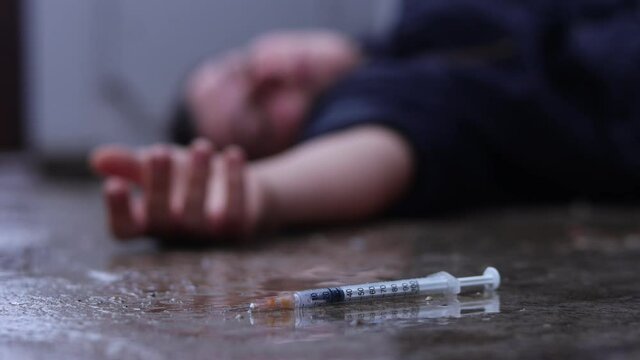 A drug addict lies with a syringe on the street in the rain.