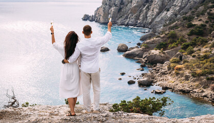 A man and a woman drink champagne on the seashore, rear view. Wedding trip to the sea, panorama. A couple in love in white clothes on the coast. Couple with champagne by the sea. Wedding travel - Powered by Adobe