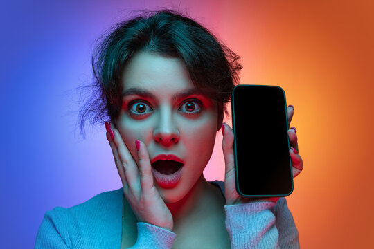 Close-up of shocked, surprised young pretty girl in warm gray knitted sweater with phone isolated on gradient blue orange neon background