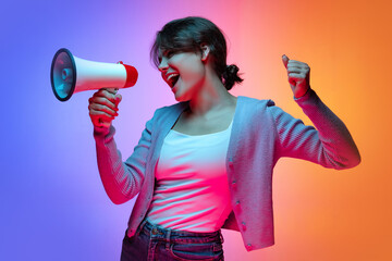 Excited young beautiful girl in warm cardigan shouting at megaphone isolated on gradient blue...