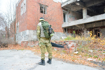 A stalker in overalls with a gas mask and a backpack stands at an abandoned building. A soldier in special equipment at the ruins of a house. 