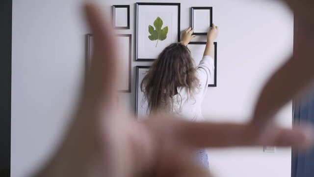 Couple arrangements wall in apartment Person holds fingers in photo frame assist lady with long loose curly hair decorating wall with pictures of different size in new flat