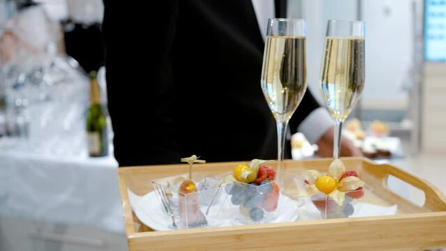 A waiter in a tailcoat demonstrates a tray with two glasses of sparkling wine or champagne and a fruit snack.