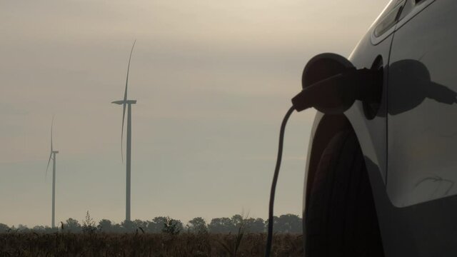 Wind turbines prodduce electricity on cloudy weather. Modern electric car charged by clean power against turbines rotating in field close view