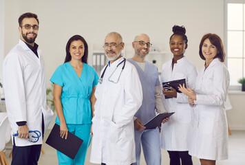 Diverse clinic staff at work. Team of happy young and mature doctors, clinicians, therapists, cardiologists in scrubs and white coat uniforms standing in office, holding clipboards, looking at camera