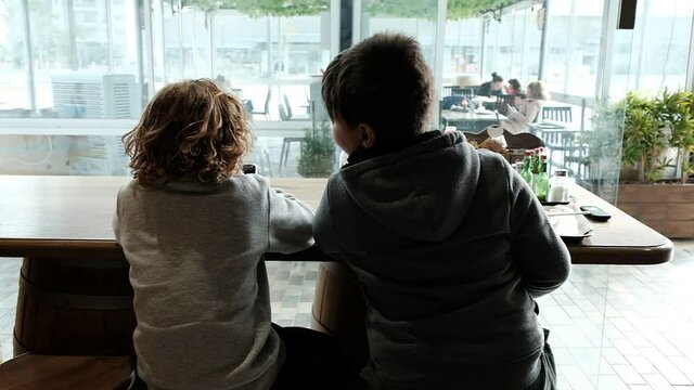 Two boys, taken from their back and not recognized, are sitting by a tall restaurant table and playing with a mobile device. Arad, Israel.