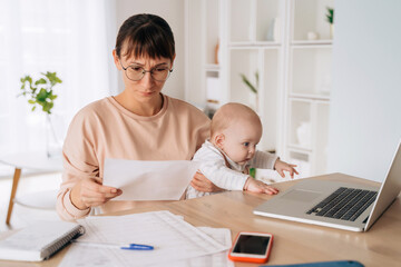 Young single mother brunette holding her toddler while trying to solve financial problems, looking...