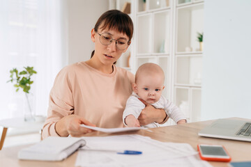 Beautiful serious business mom trying to work while spending time with her cute baby boy at home. Young working mother looking sadly at documents, sitting at her home office with her infant baby