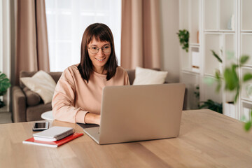Pleasant happy young woman freelancer working on laptop at home. Attractive businesswoman in eyeglasses studying online, using laptop software, web surfing information or doing online shopping