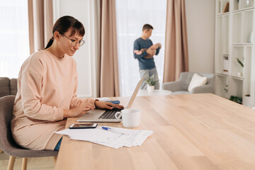 Young woman wife and mother remote working online from home office while her husband taking care of their infant baby. Man in paternity leave instead his working wife. Busy mom working with papers