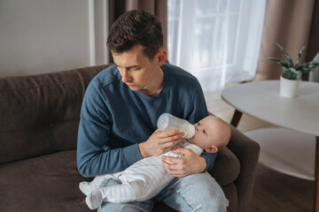 Unhappy young father feeding newborn baby with milk bottle on couch at home. Depressed single dad tired from sleepless night, fatherhood, take care of infant sun. Paternity and paternity leave concept - obrazy, fototapety, plakaty