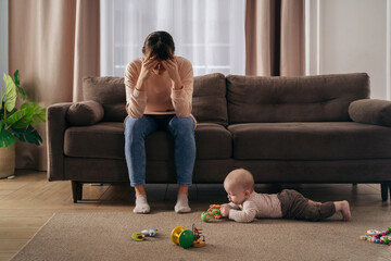 Young single frustrated woman hold her head with hands sitting on sofa in living room while her toddler son playing with toys on floor. Exhausted mom suffering from postpartum post-natal depression  - obrazy, fototapety, plakaty