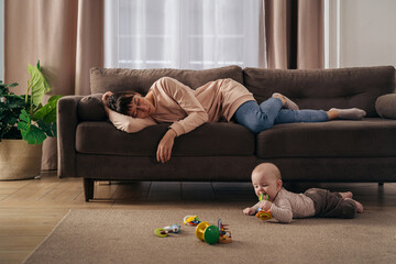 Young tired mother suffering from lack of sleep, sleeping on sofa while her little infant baby is...