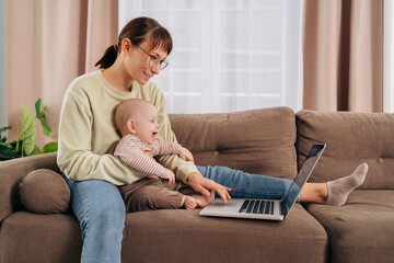 Happy young woman working from home with her adorable cheerful baby, sitting on couch, using...