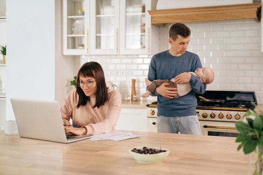 Young Mother Working Remotely From Home While Her Husband Babysitting With Their Little Child. Woman In Glasses Using Laptop In Kitchen While Man Holding Their Sleeping Toddler Baby. Paternity Leave