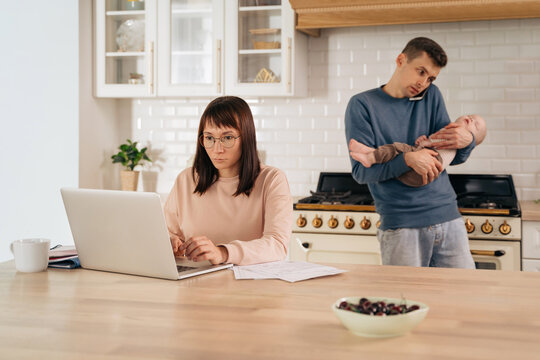 Paternity Leave And Gender Family Roles Concept. Young Business Woman Using Laptop For Remote Work From Home While Her Multitasking Husband Take Care Of Their Toddler Infant Baby And Taking Phone Call