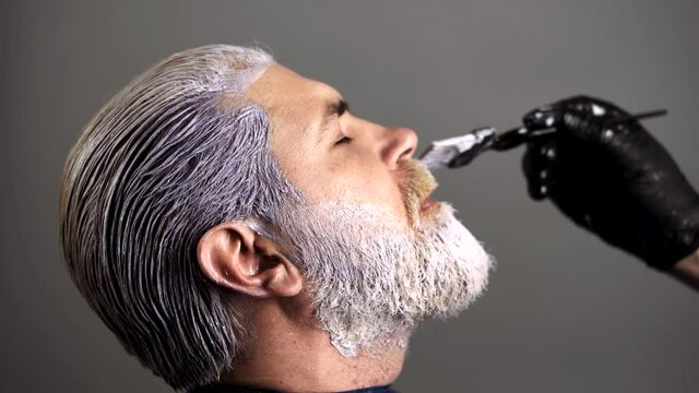 Process of a guy having beard and moustache dyed at hairdresser salon. Hipster bearded men dye his hair color on a gray. Hairdresser colorist dyes the mustache hair of a bearded man.