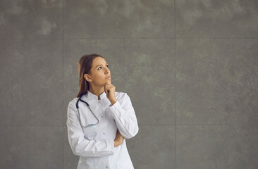 Pensive young female doctor holding her chin imagining or thinking about something important. Doctor stands on a gray background and directs his gaze to a free space for text. Medical issue concept.