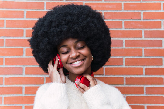 Portrait of beautiful african american woman with eyes closed, white perfect smile, long afro hair and white jumper listening to music with red headphones in brick wall. Copy space for text