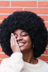 Portrait of cheerful african american woman with white perfect smile, long afro hair and white jumper covering eye against brick wall. Beautiful afro woman with eyes closed. Dentist concept