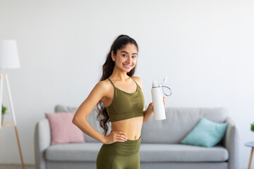 Millennial Indian woman in sportswear drinking water or protein shake from bottle at home