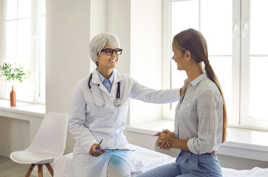 Smiling friendly female doctor touches patient's shoulder assuring that everything will be fine. Doctor sets young woman positive during conversations in hospital room. Concept of medical support.