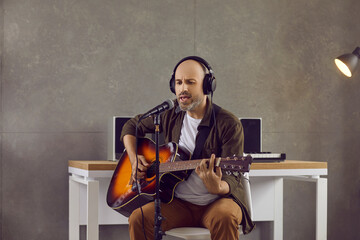 Bald adult man in headphones sitting on chair, playing acoustic guitar and singing into microphone....
