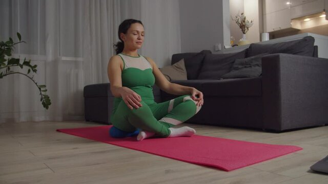 Attractive adult woman in sportswear sitting on yoga pillow and mat floor in front of open laptop indoors, keeping eyes closed, legs crossed, meditating to nature sounds using app via Internet at home