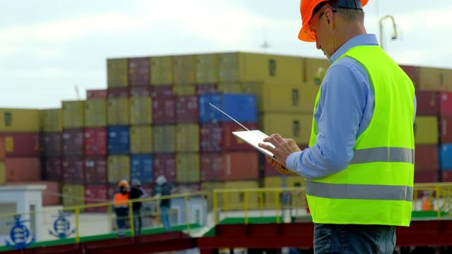 Cargo ship loading in dock. Safety manager in colorful uniform marks notes on tablet of employees work with metal containers in dockyard