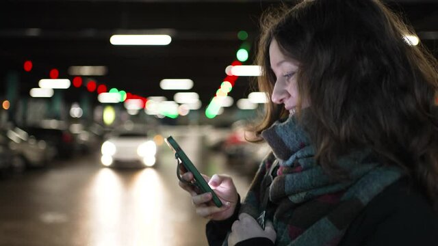 Side view of a young woman with a mobile phone in her hands walking along the parking lot of a shopping center between the rows with cars, looking at the screen and smiling, online communication