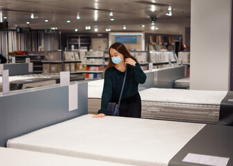 Pleasant young woman in medical face mask choosing new mattress in store during pandemic