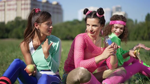 Retro friends teasing young woman eating chocolate bar outdoors sitting on sunny lawn. Portrait of fit confident Caucasian women with healthful fruits mocking lady with sweet dessert in slow motion