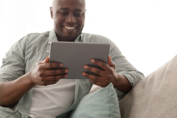 African american man using digital tablet and smiling, reading book or having video call while sitting on couch, closeup