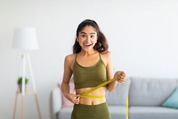 Portrait of young Indian woman measuring her waist with tape indoors, smiling at camera