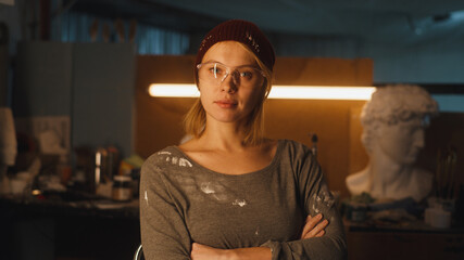 Blond craftswoman in goggles and hat looking at camera while standing against workbench with busts in dimly illuminated workshop