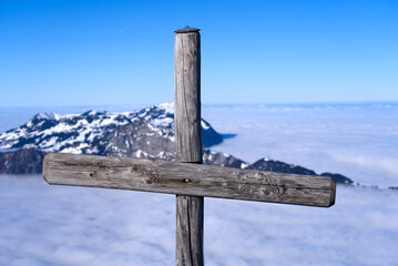 Wooden cross at mountain Fronalpstock at mountain village Stoos on a sunny winter day with sea of fog and mountain panorama. Photo taken December 20th, 2021, Stoos, Switzerland.