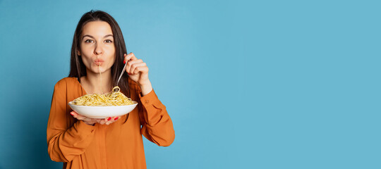 Beautiful young girl eating delicious Italian pasta isolated on blue studio background. World pasta...