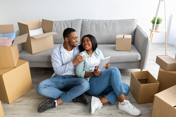 Excited black spouses with touch pad searching household goods for their new home, using tablet on moving day