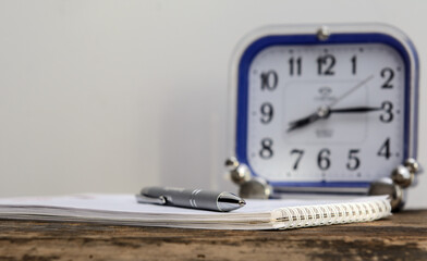 There is a ballpoint pen on a notebook lying on an old blackboard. There is an alarm clock in the background. Concept business