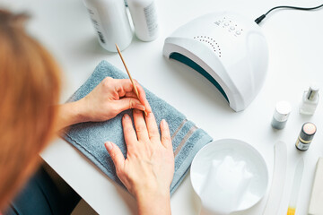 Woman preparing nails to apply gel hybrid polish using UV lamp. Beauty wellness spa treatment concept. Cosmetic products, UV lamp, green leaves on white table. Spa, manicure, skin care concept